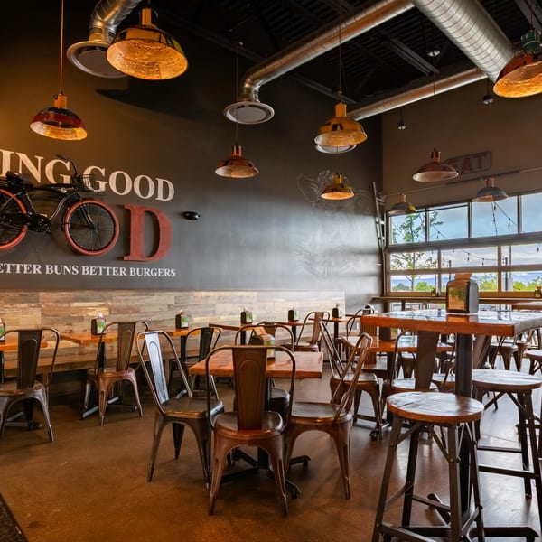 Interior of Skirted Heifer restaurant with industrial lighting, wooden tables, metal chairs, and large windows.