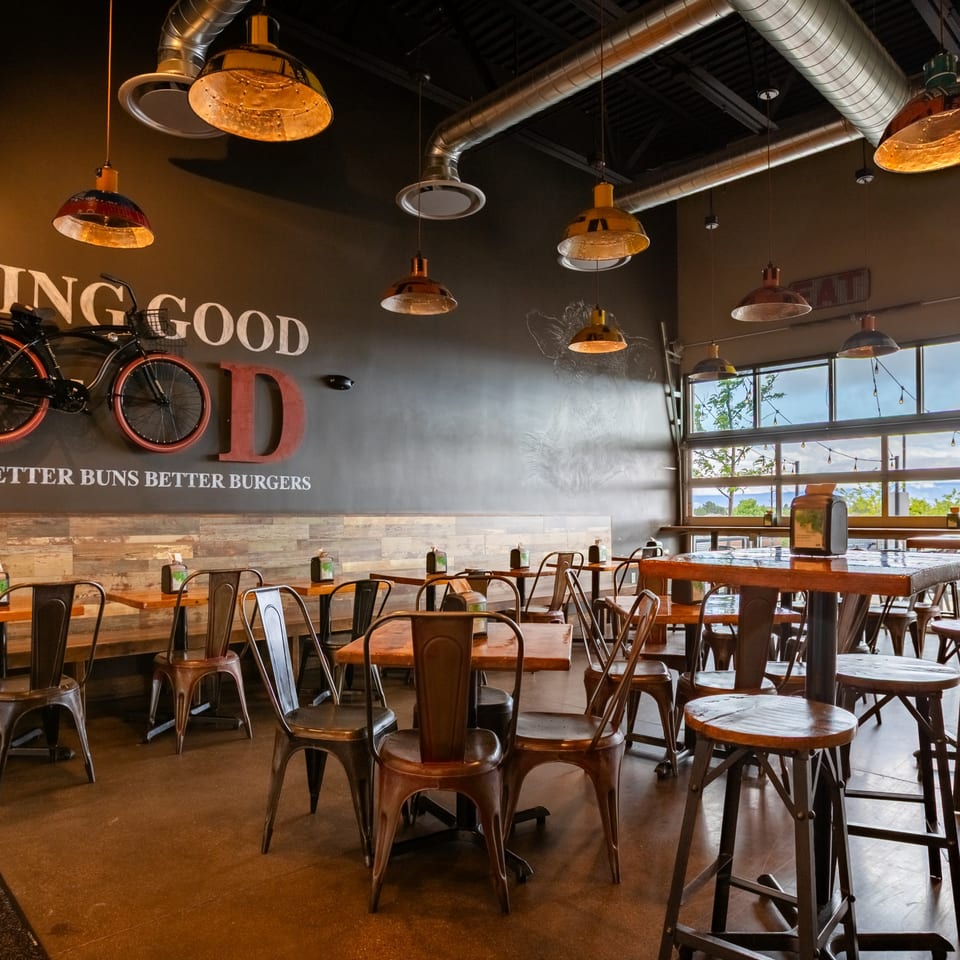 Interior of Skirted Heifer restaurant with industrial lighting, wooden tables, metal chairs, and large windows.