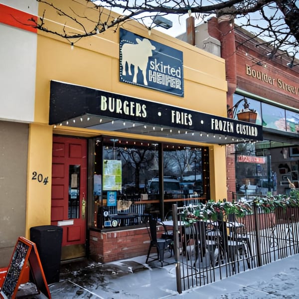 The Skirted Heifer burger restaurant exterior on a snowy day, with outdoor seating and a red door.