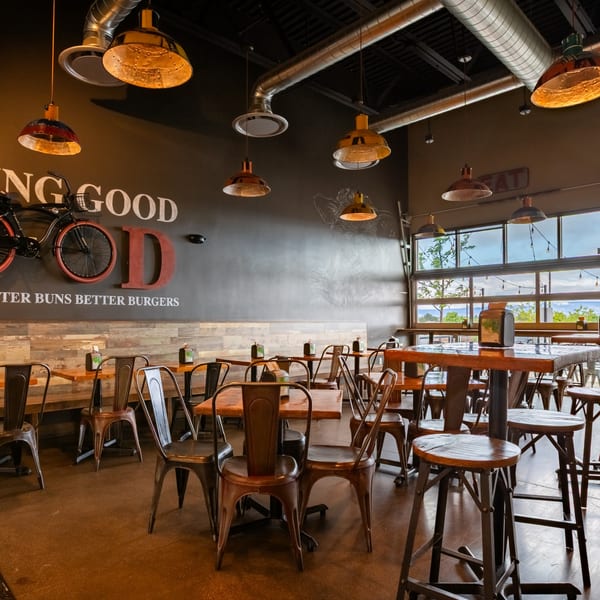 Interior of Skirted Heifer restaurant with industrial lighting, wooden tables, metal chairs, and large windows.