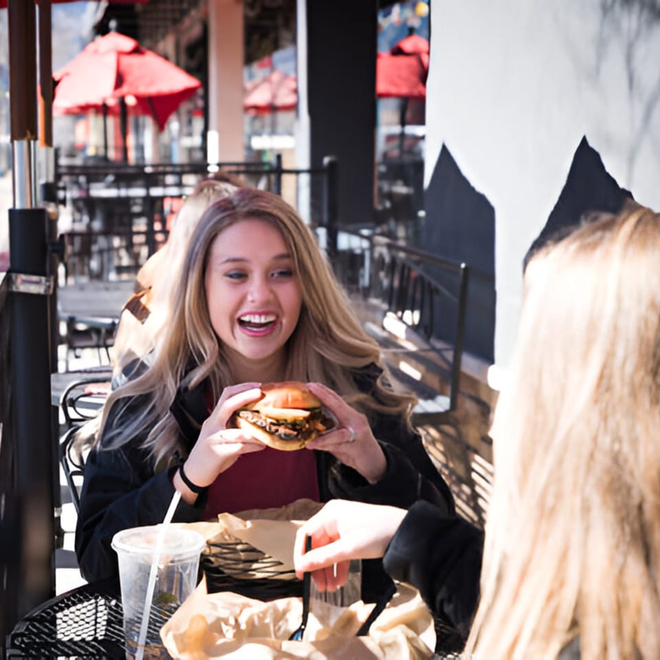 A smiling woman holding a Skirted Heifer burger, dining outdoors with a friend.