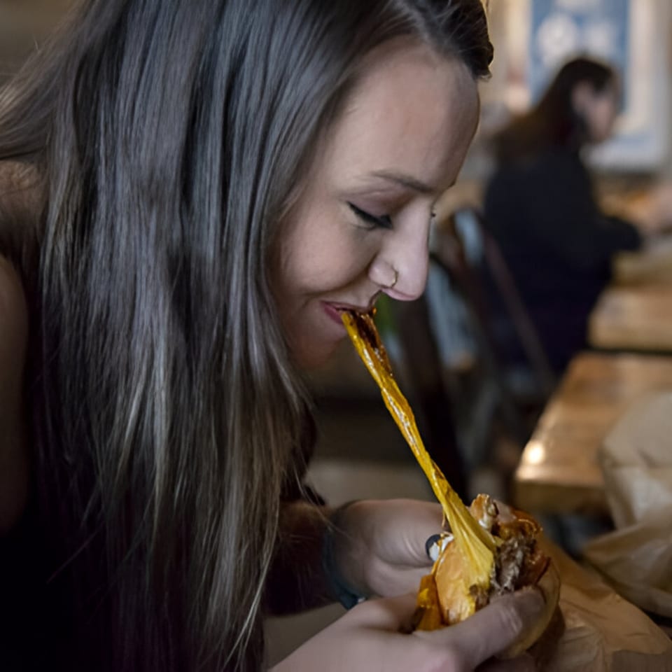A woman happily eating a Skirted Heifer burger with melted cheese stretching from her mouth.