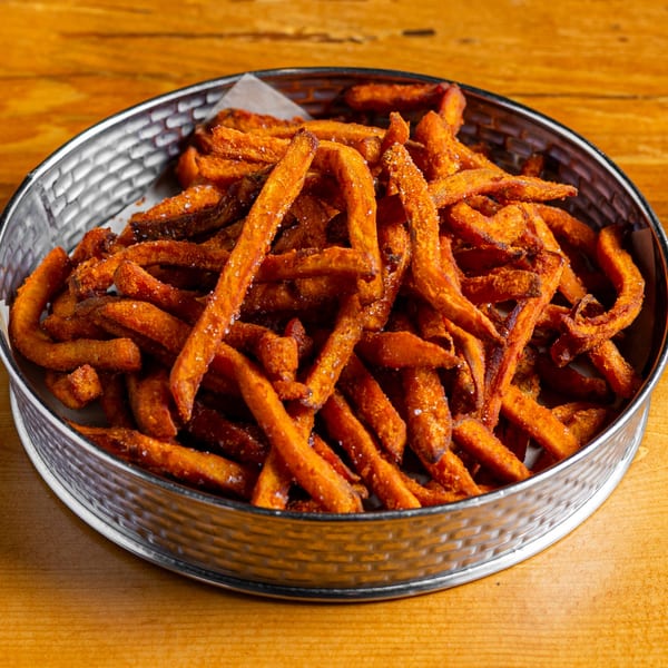A metal basket filled with crispy, golden-orange sweet potato fries, lightly salted, on a wooden table.