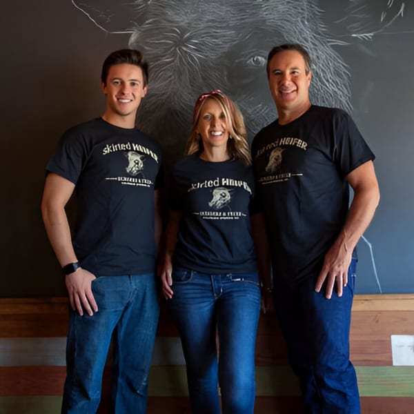 Three smiling Skirted Heifer staff members in branded t-shirts stand before a chalk cow drawing.