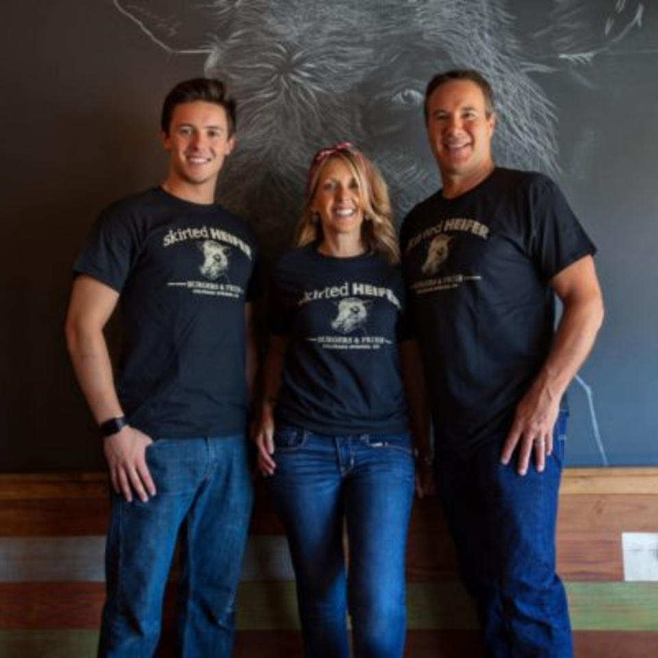 Three smiling Skirted Heifer staff members in branded t-shirts stand before a chalkboard cow drawing.