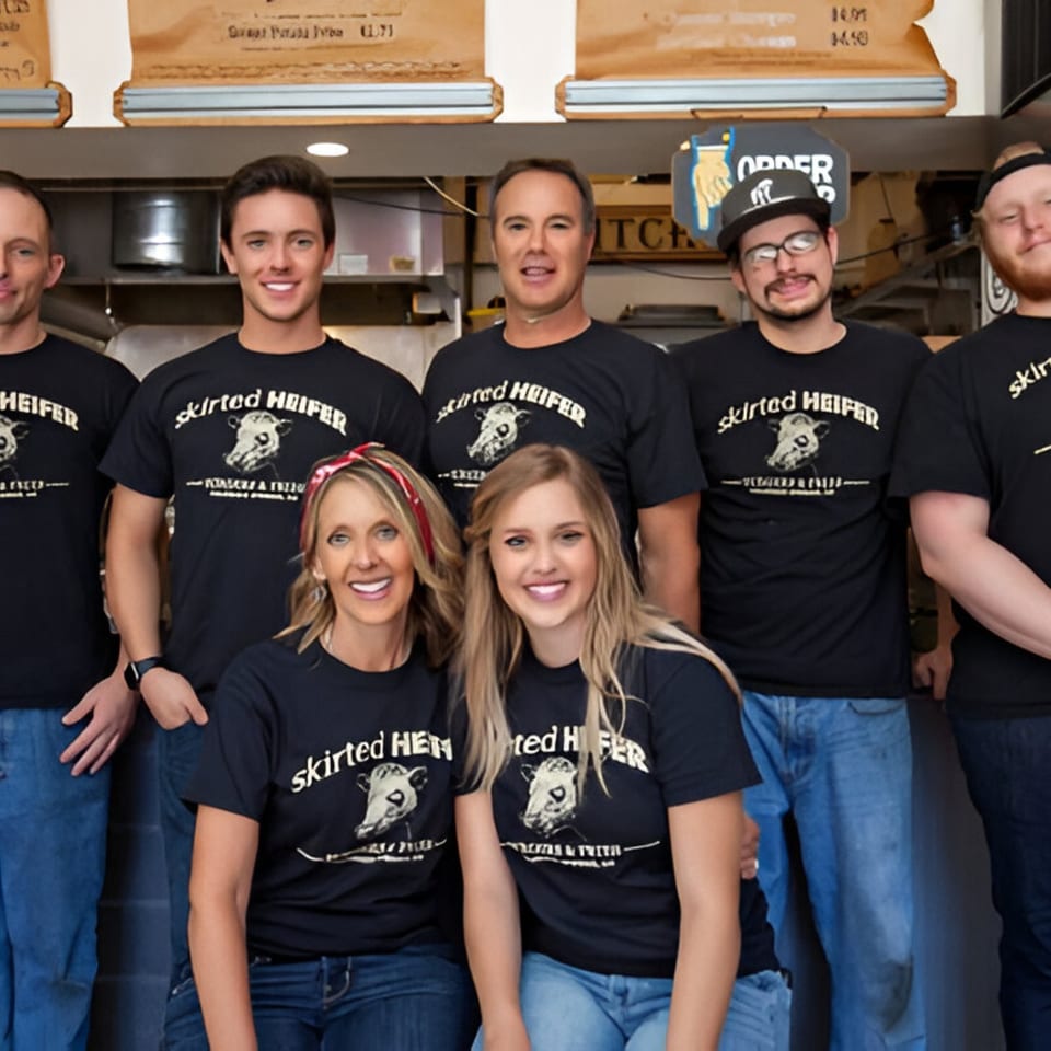 Six smiling Skirted Heifer staff members in black t-shirts and jeans pose together.