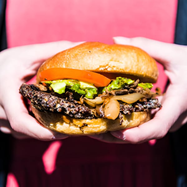 Hands holding a Skirted Heifer burger with a dark patty, lettuce, tomato, and caramelized onions.