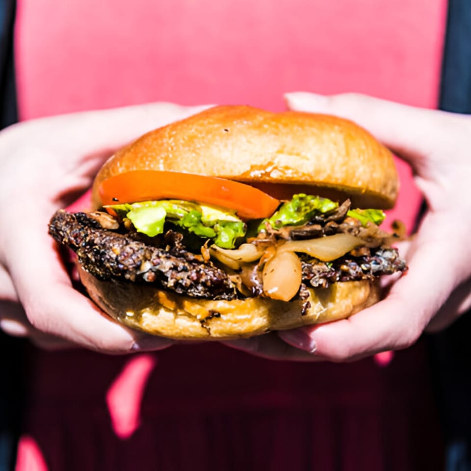 Hands holding a Skirted Heifer burger with a dark patty, lettuce, tomato, and caramelized onions.