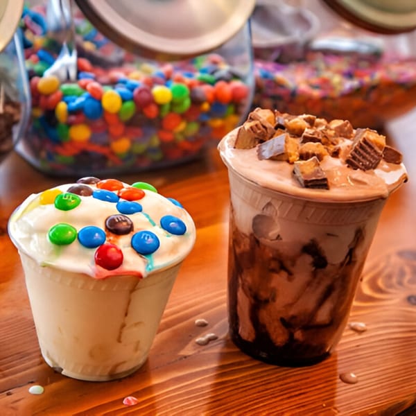 Two Skirted Heifer milkshakes, one with M&Ms and one with Reese's, on a wooden table.