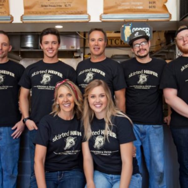Seven smiling Skirted Heifer staff members in black logo t-shirts pose together in the restaurant.