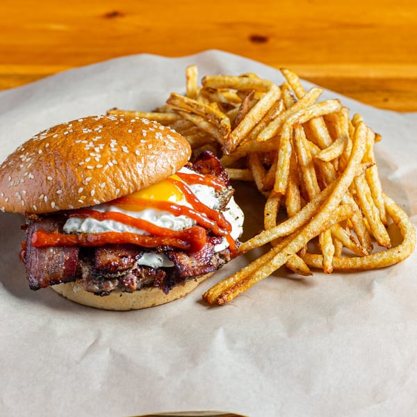 A Skirted Heifer burger with bacon, fried egg, and ketchup, served with a side of fries.